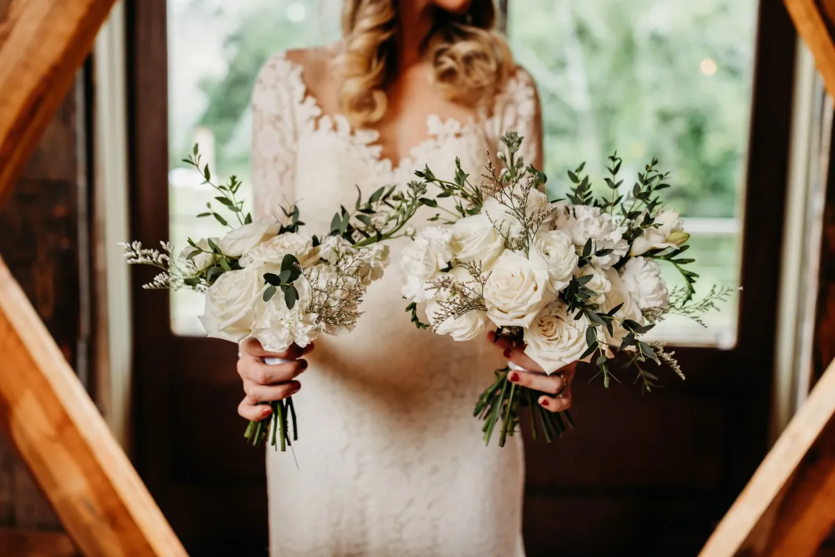 Bride holding two lush white bouquets in a wooden arch
